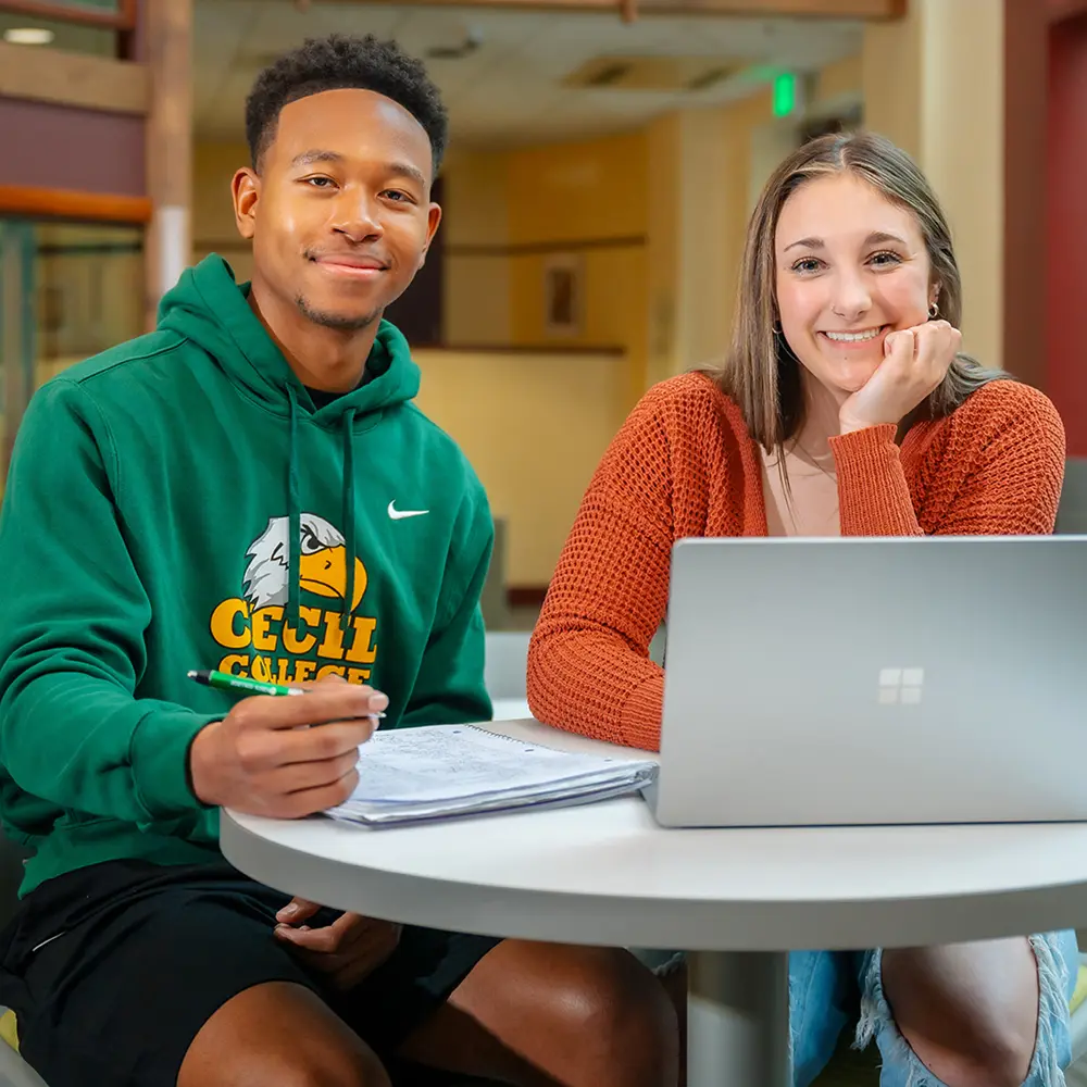 Two Cecil College students sitting at a table in the student lounge with laptop, smiling at the camera. fall clothing.