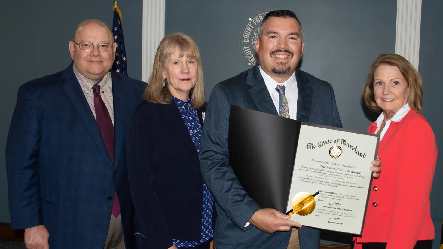 The image shows four people standing together in a formal setting, likely for an award or recognition ceremony. On the left, a man in a dark blue plaid suit with a burgundy tie stands with his hands at his sides. Next to him, a woman with shoulder-length blonde hair is wearing a navy blue blazer over a patterned blouse. To her right, a man holding a certificate smiles broadly; he is dressed in a dark suit with a white shirt and patterned tie. The certificate is prominently displayed, featuring a gold seal. On the far right, another woman wearing a coral blazer over a white top is smiling. The background features a deep blue wall with a decorative emblem and an American flag partially visible.